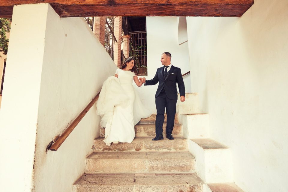 Una pareja de recién casados baja de la mano por una escalera de piedra en un edificio de paredes blancas.