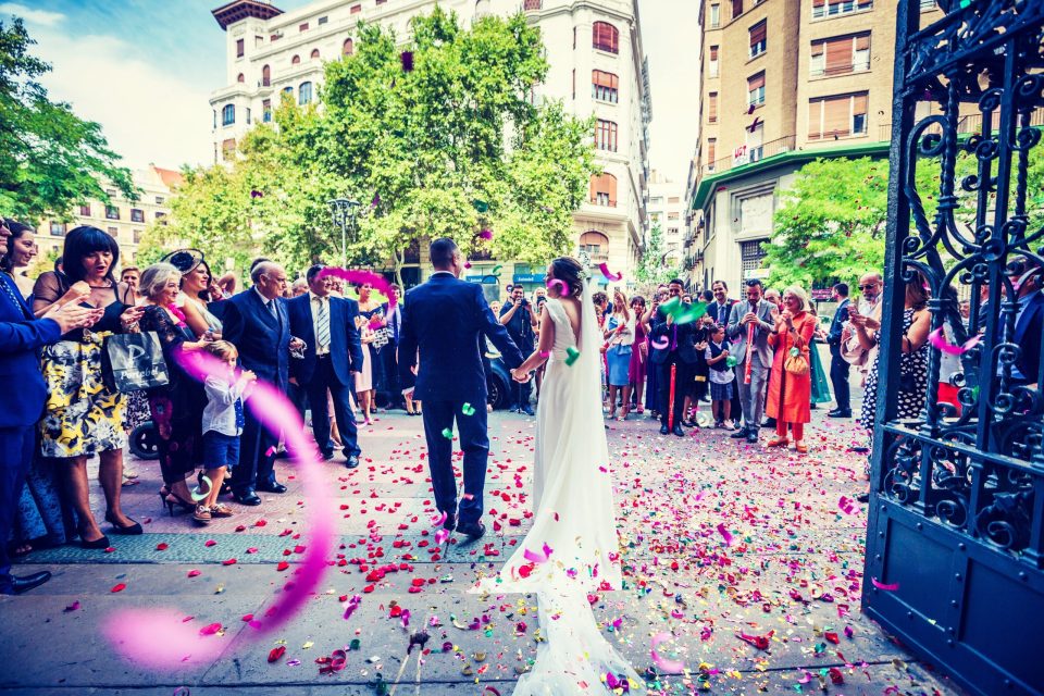 Una pareja de recién casados camina frente a la entrada de una iglesia mientras familiares e invitados los reciben y lanzan.