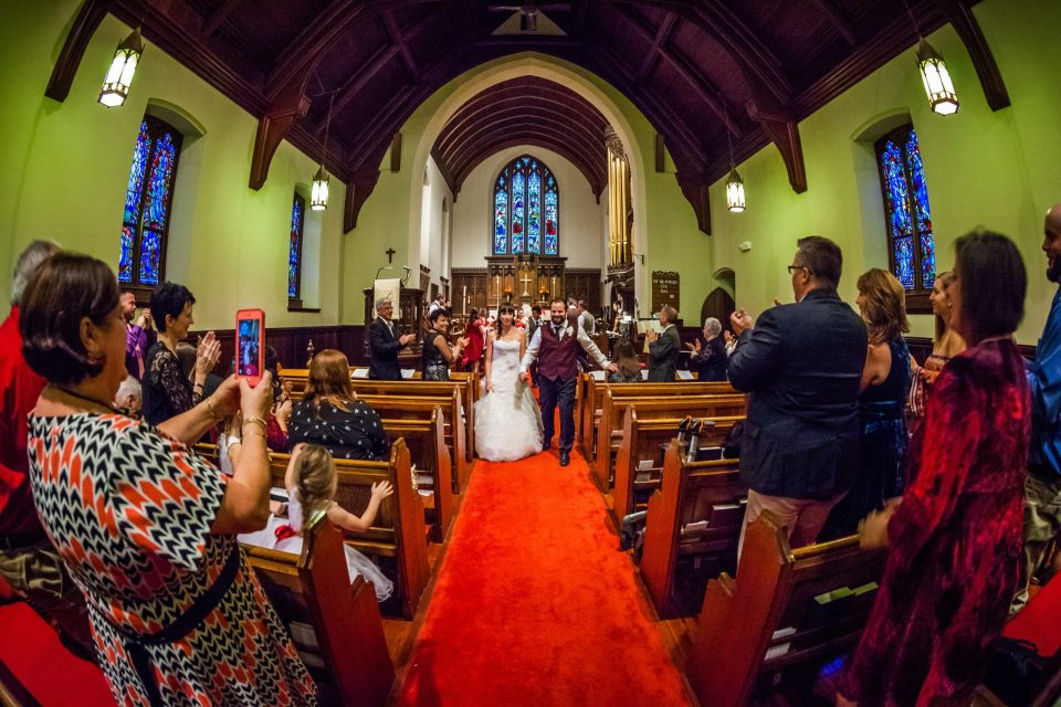 A newlywed couple walks down the central aisle of a church on a red carpet.