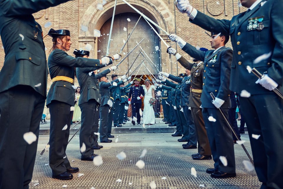 Una pareja de recién casados sale de una iglesia mientras atraviesa un arco ceremonial formado por personas uniformadas.