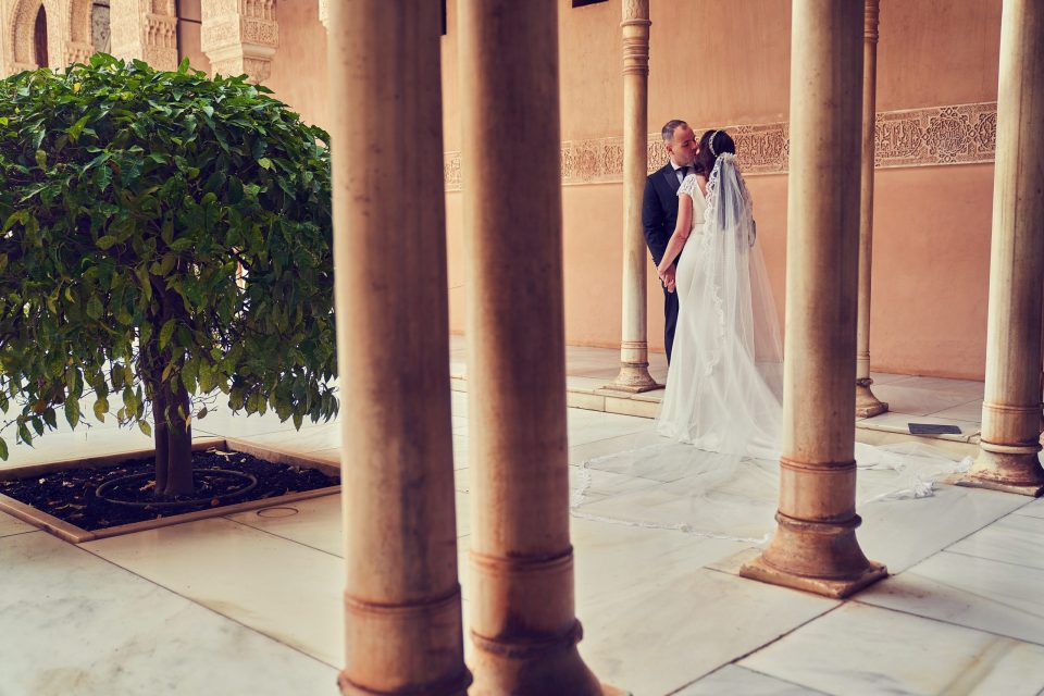 Una pareja de recién casados se besa bajo una galería de columnas, en un patio con arquitectura ornamental.