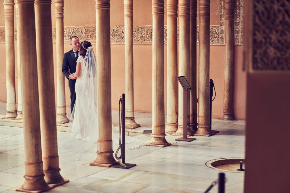 Una pareja de recién casados se mira entre una fila de columnas ornamentadas en un patio de arquitectura decorativa.
