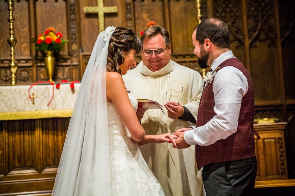 A couple stand together, with the woman wearing a wedding dress and veil and the man wearing a burgundy waistcoat.
