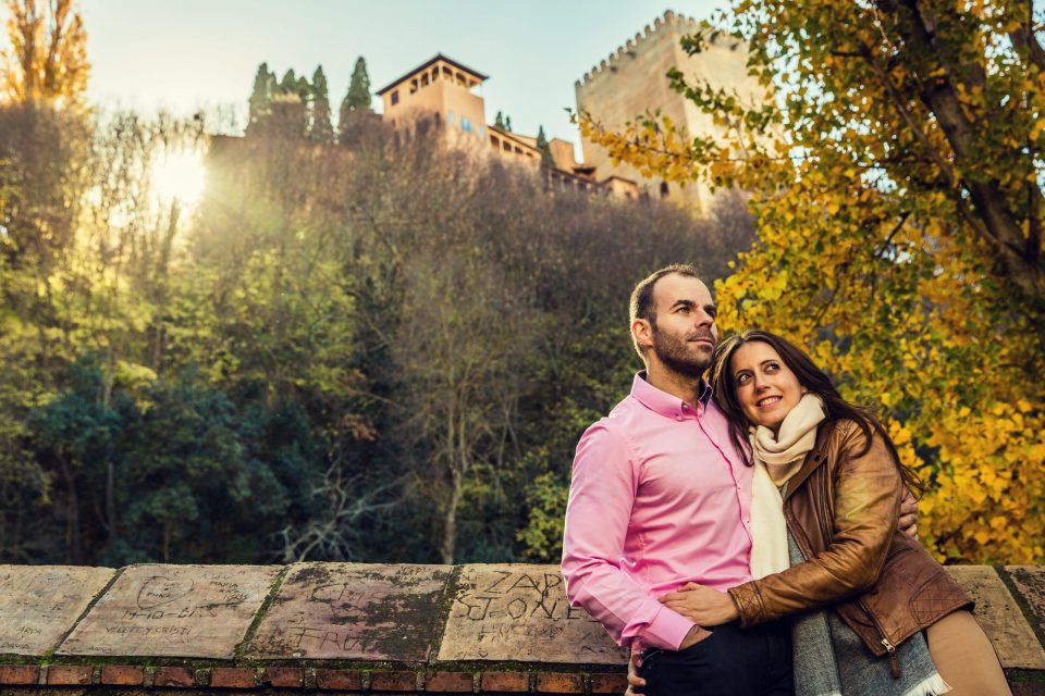 Una pareja posa abrazada en el exterior, apoyada sobre un muro de piedra con inscripciones.