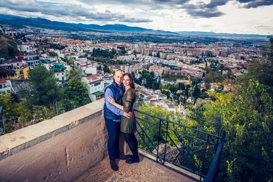 A couple pose embracing at a lookout with a railing.