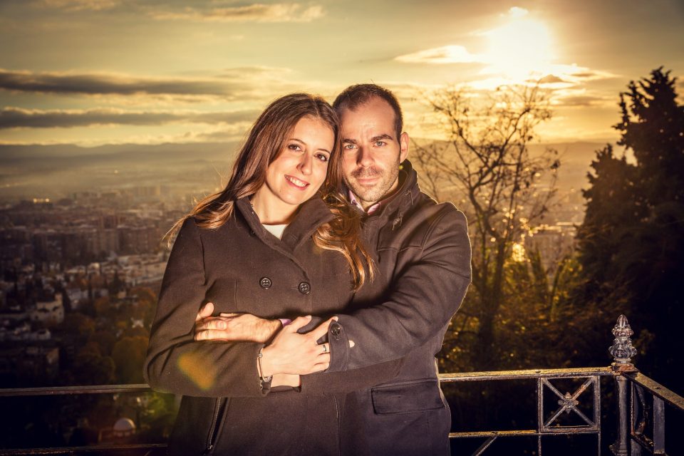 A couple pose embracing in front of a railing, with a city and trees in the background.