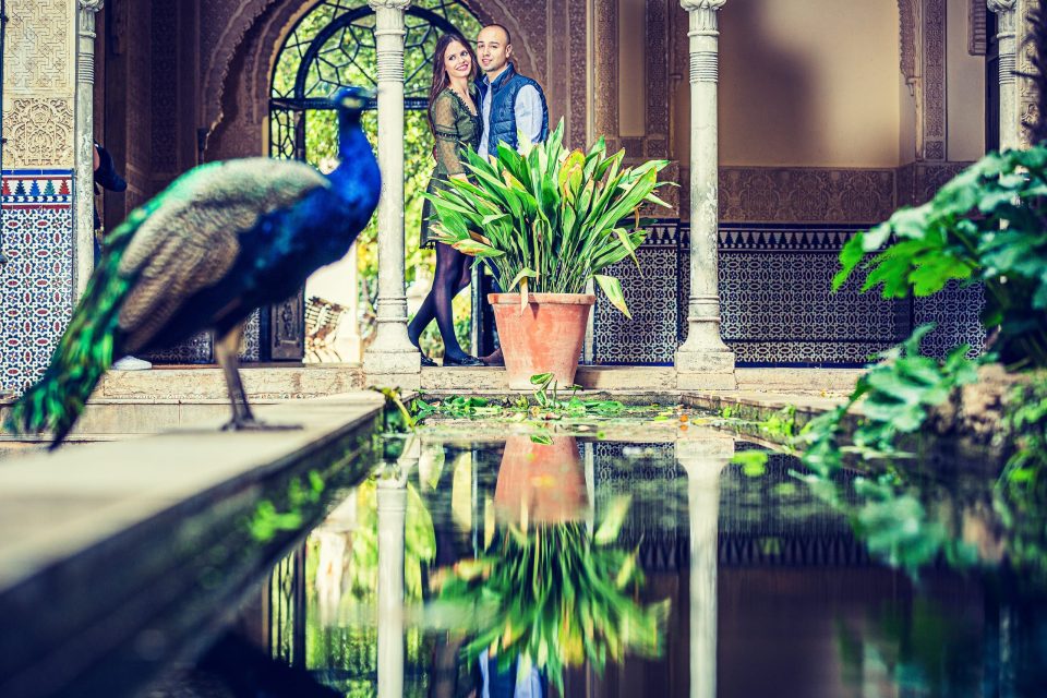 Una pareja posa bajo una galería con columnas, azulejos ornamentales y un arco decorado.