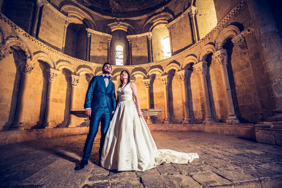A couple poses inside a church or historic stone building, beneath a dome with arches and decorated columns.