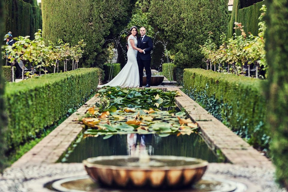 Una pareja posa en un jardín formal, entre setos recortados y cipreses.