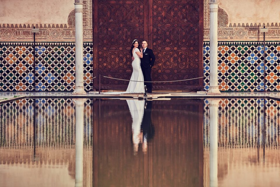 Una pareja posa frente a una gran puerta de madera tallada, entre columnas y paredes decoradas con azulejos geométricos.