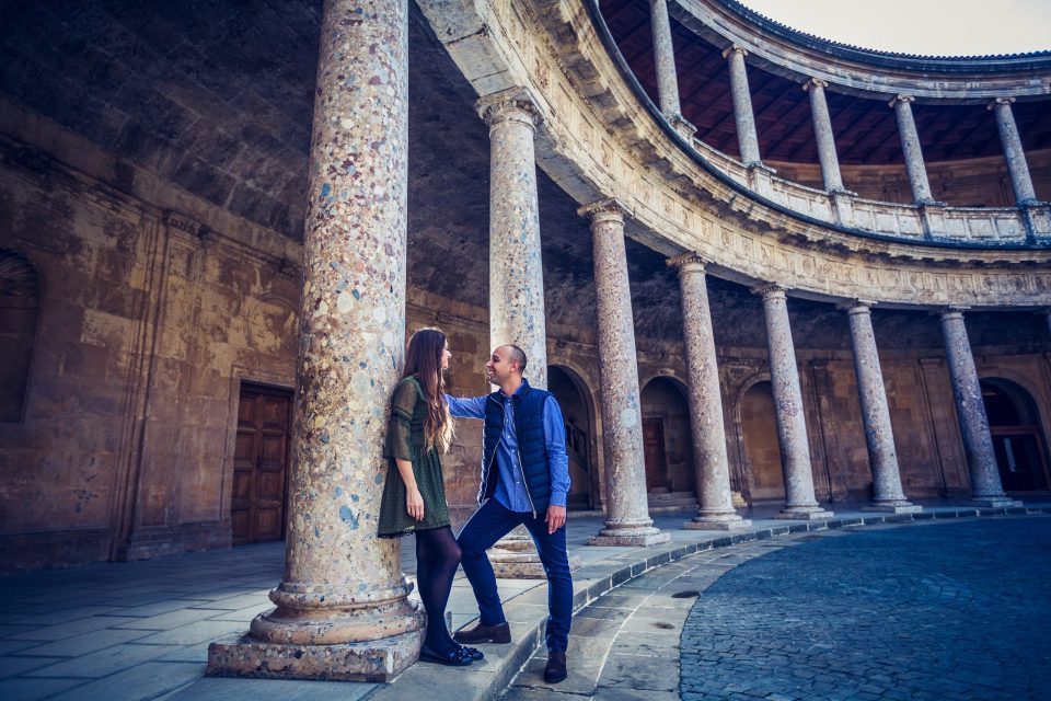 A couple pose together in the semicircular courtyard of a historic building, surrounded by stone columns.