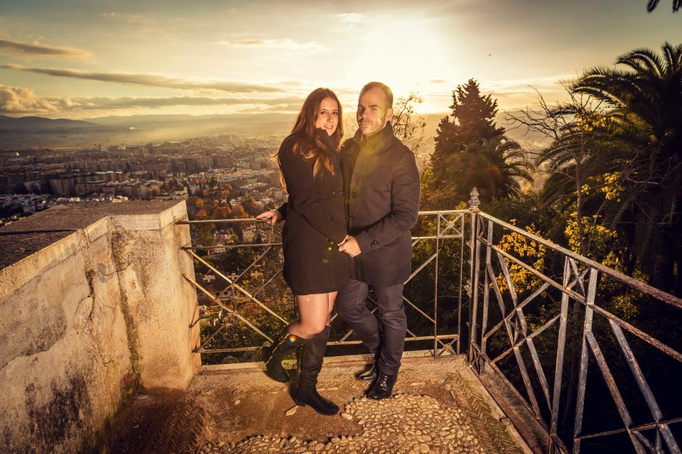 A couple poses together on an elevated terrace with an iron railing, overlooking a sweeping urban and mountainous panorama.