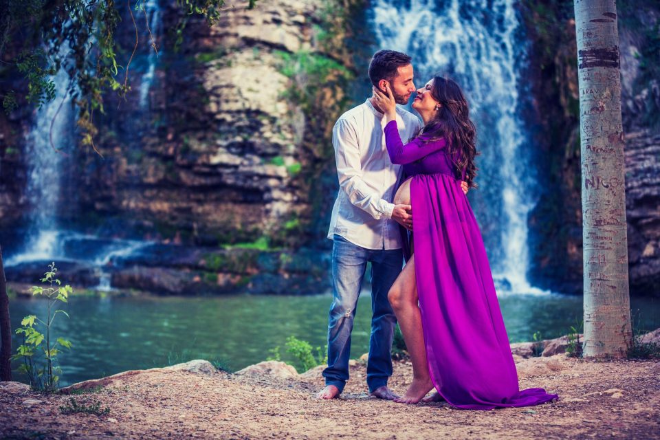 Una pareja posa junto a una cascada y una pequeña laguna en un entorno arbolado.