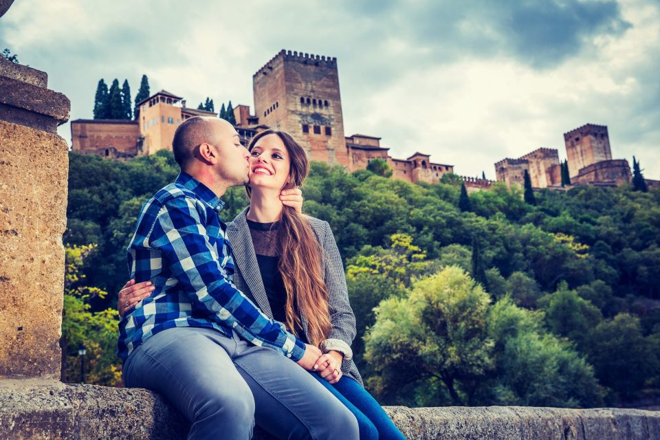 Una pareja posa sentada en un muro de piedra mientras una persona besa suavemente la mejilla de la otra.