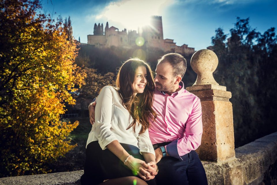 A couple pose seated beside a stone balustrade, with a fortified historic building in the background.