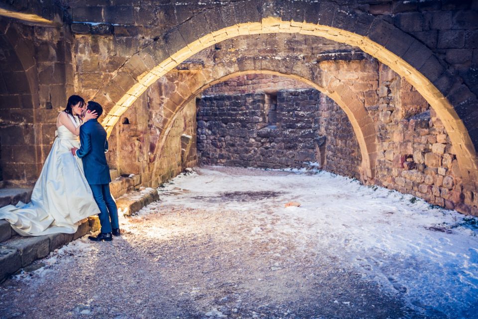 A couple embrace and kiss beside an ancient stone arch in a snow-covered setting.