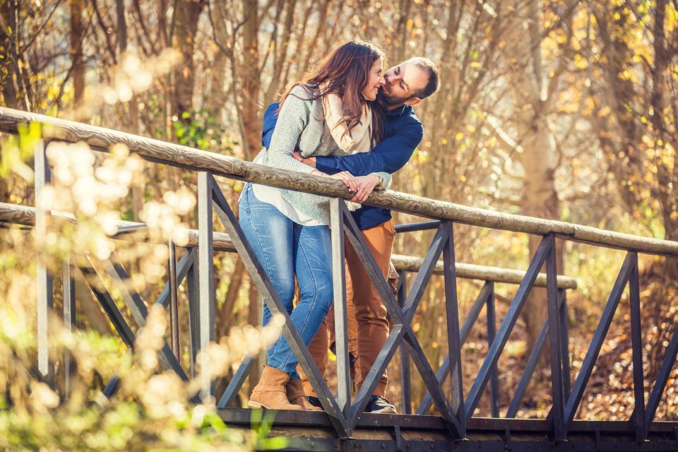 A couple embraces and kisses on a wooden bridge with metal railings.