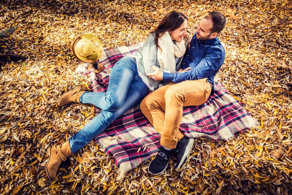 A couple embrace and look at each other while lying on a checked blanket among dry leaves.