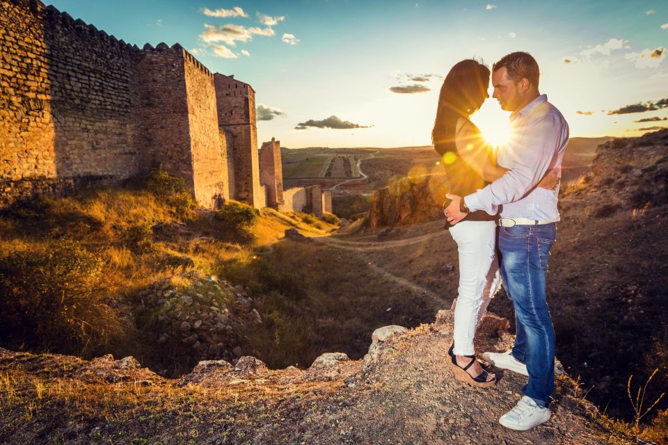 Una pareja se abraza y se mira sobre un terreno rocoso junto a una antigua fortificación de piedra.