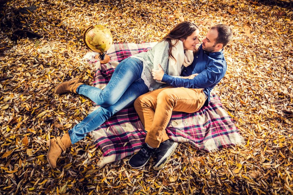 A couple embrace and look at each other on a checked blanket among dry leaves.