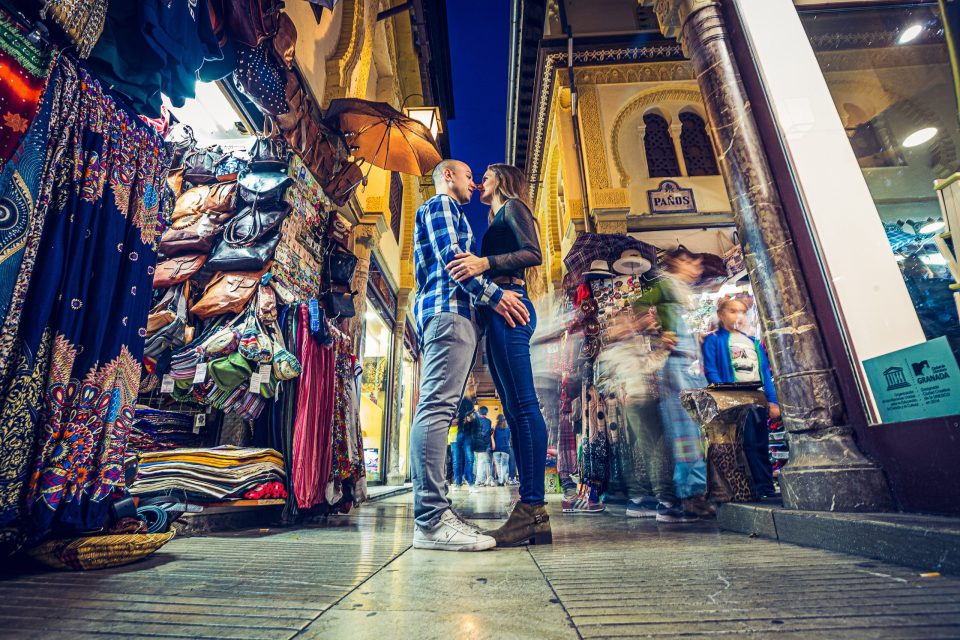 A couple kiss inside a covered market or shopping street, surrounded by fabric stalls.