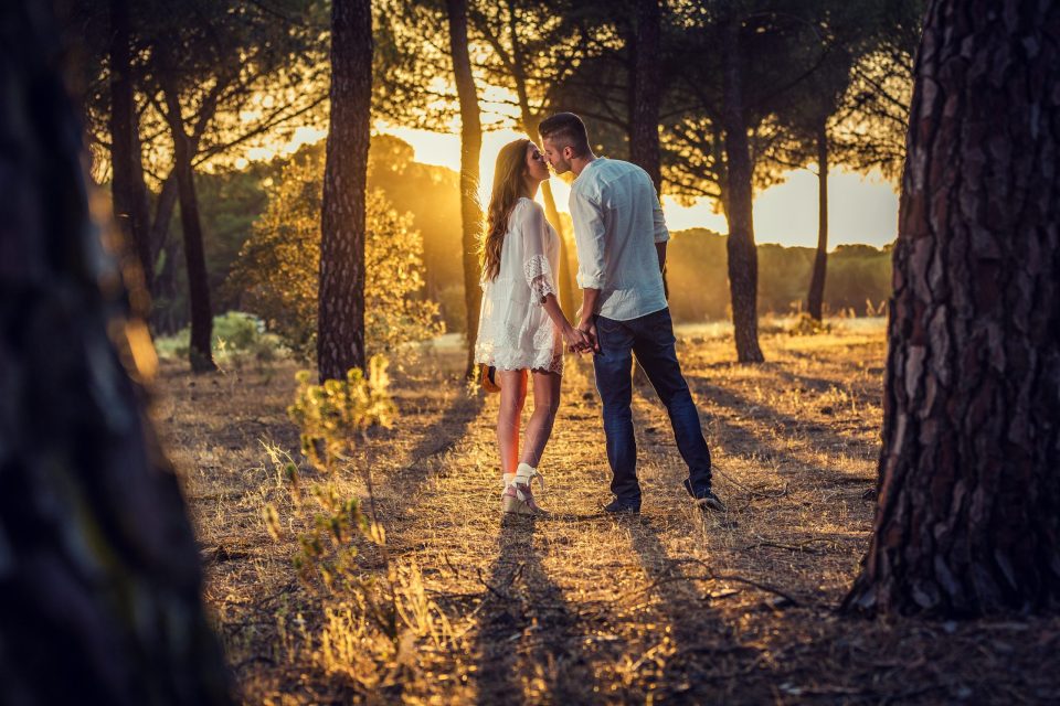 Una pareja se besa y se toma de la mano entre árboles en un campo, iluminada por la cálida luz del atardecer a contraluz.
