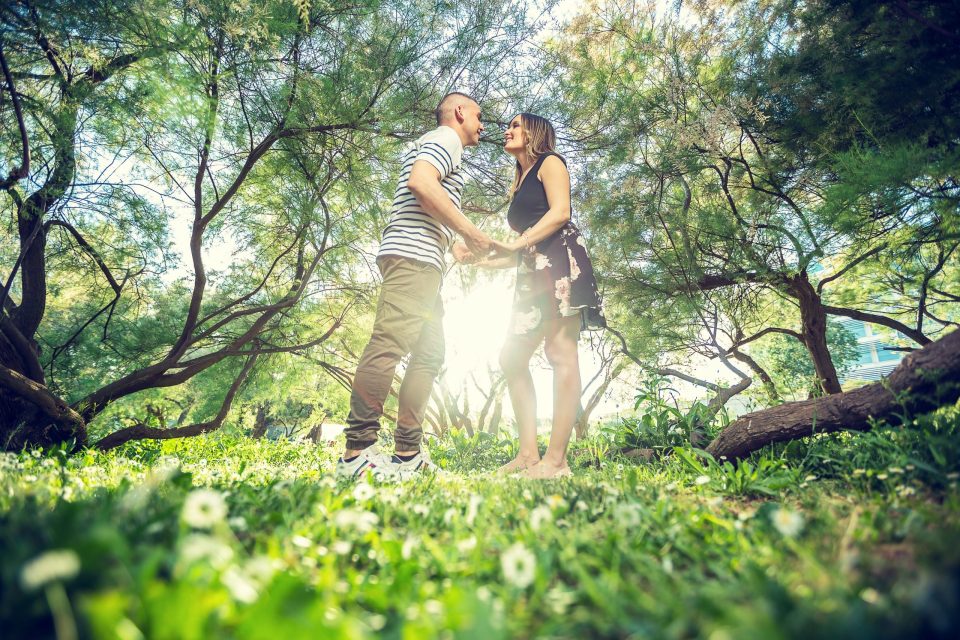 Una pareja se toma de las manos y se mira en un parque arbolado, rodeada de vegetación y pequeñas flores blancas.
