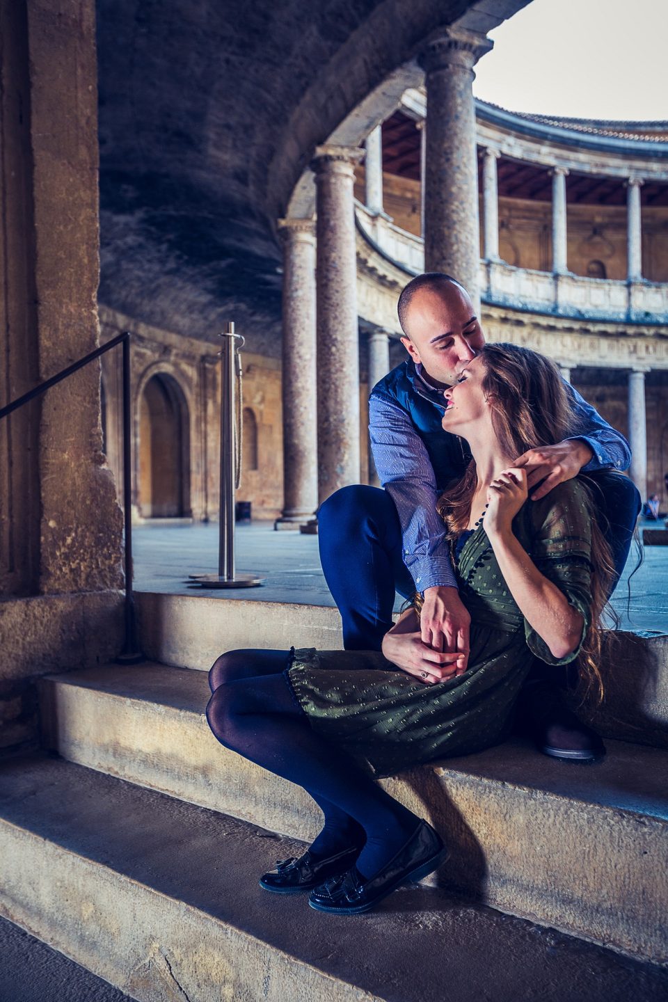 A couple sitting on stone steps embrace and kiss beneath an archway.