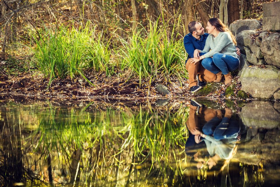 A couple sit beside rocks at the edge of a pond surrounded by vegetation and trees.