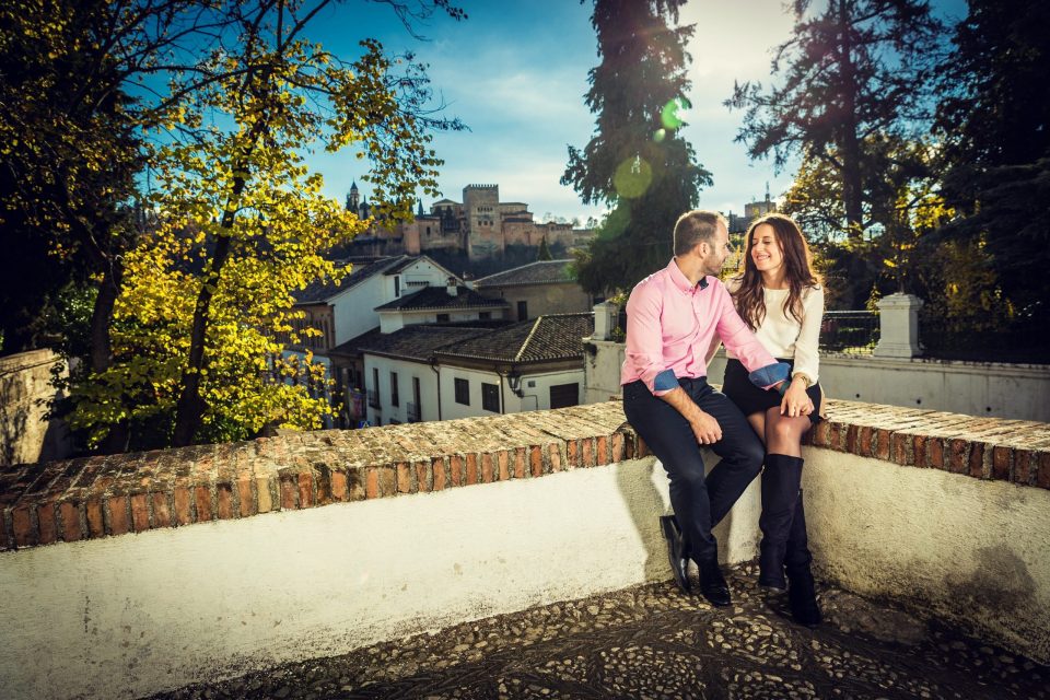 A couple is sitting on a brick wall in a sunny, wooded setting.