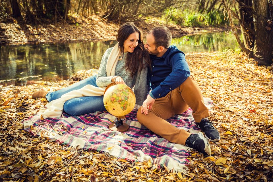 A couple sit on a checked blanket beside a pond, surrounded by trees and dry leaves.