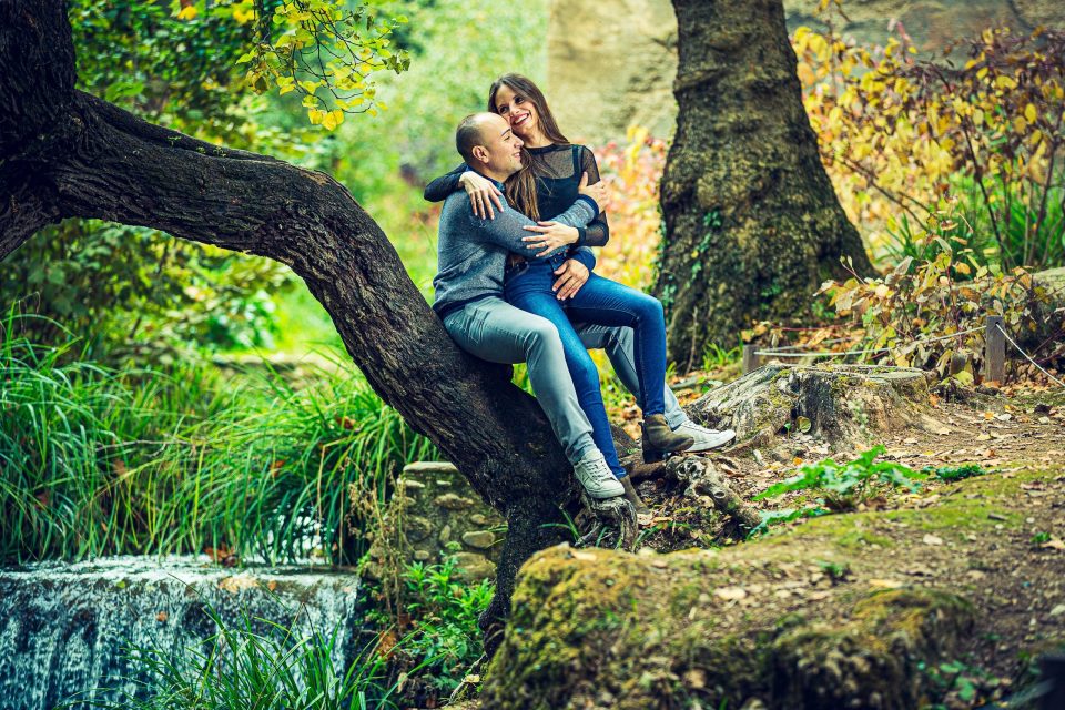 A couple smile and embrace while sitting on a sloping branch, surrounded by trees and autumn vegetation.