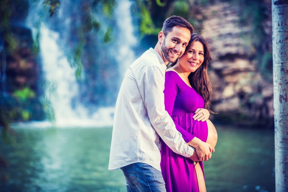 Una pareja sonriente posa junto a una cascada en un entorno natural con agua y vegetación.