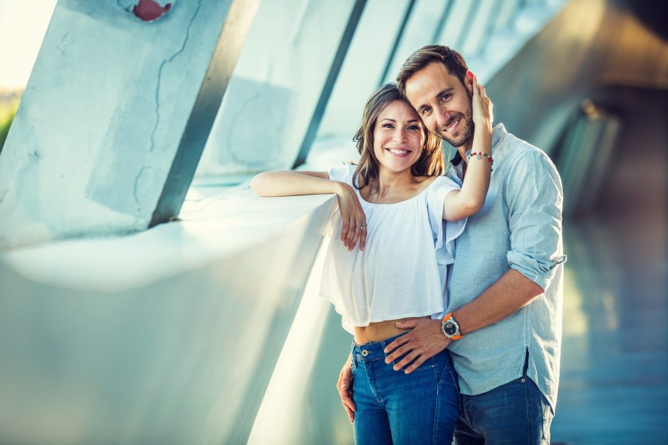 Una pareja sonriente posa muy cerca en una pasarela urbana con grandes ventanales y estructuras inclinadas.