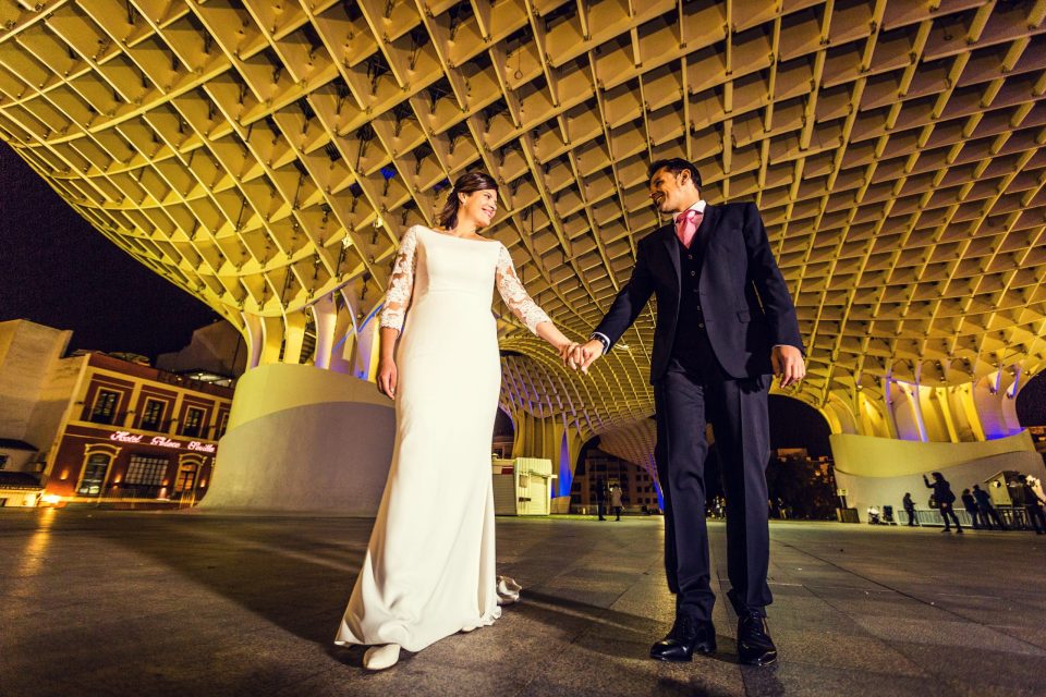 A couple in wedding attire walk hand in hand through an illuminated square at night.