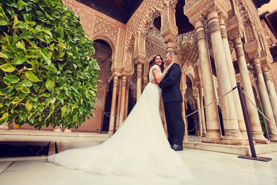 Una pareja vestida de boda posa abrazada en un patio ornamentado, rodeada de columnas.