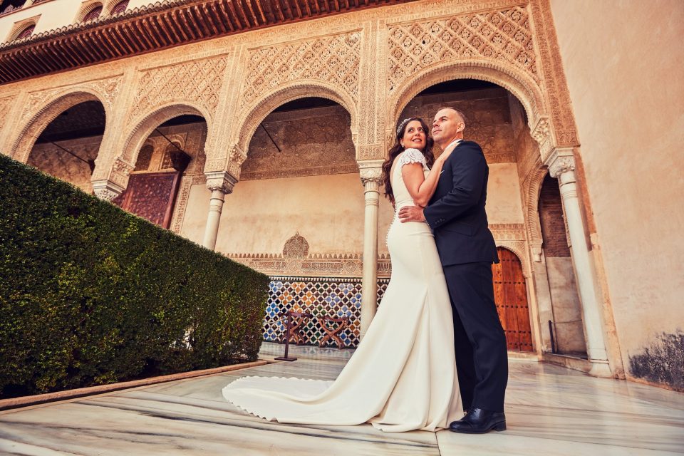 Una pareja vestida de boda posa abrazada en un patio de arquitectura ornamentada, con arcos decorados, azulejos.