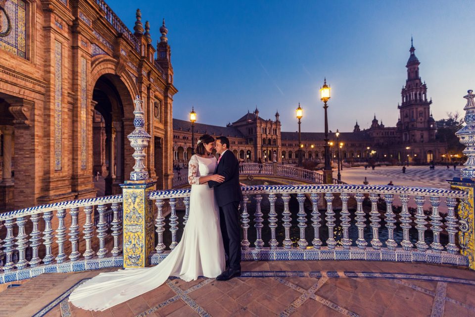 A couple dressed in wedding attire pose embracing on a tiled bridge.