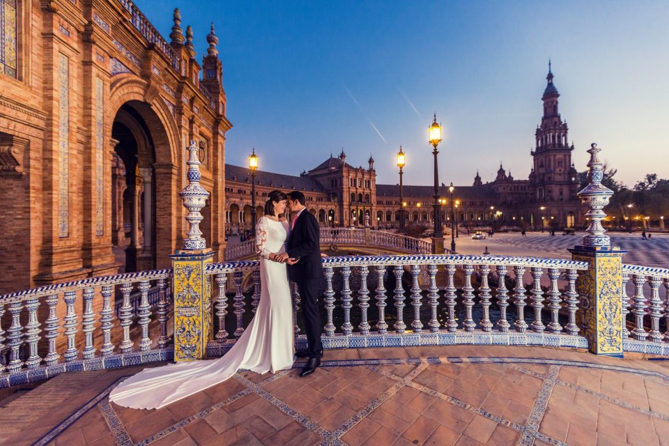 A couple dressed in wedding attire poses together at sunset on an ornate tiled balustrade.