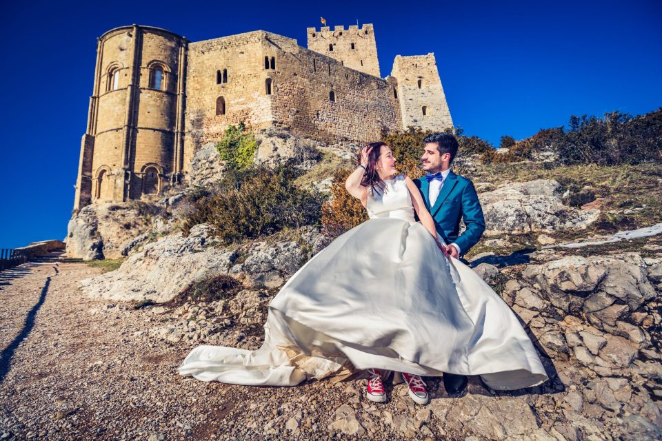 A couple dressed for their wedding pose on rocky ground in front of a stone castle.