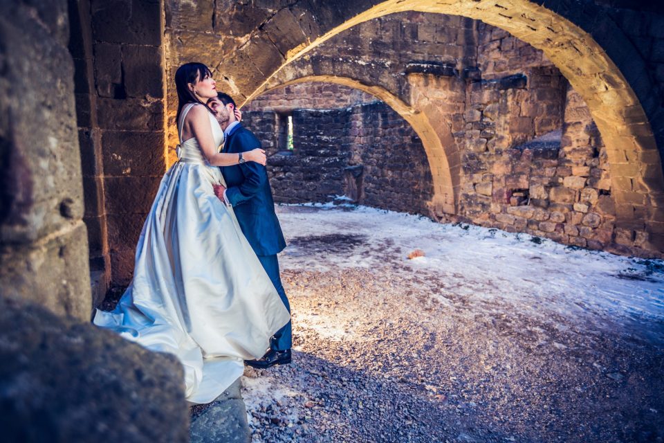 A wedding couple embrace beneath a stone arch inside an ancient building.
