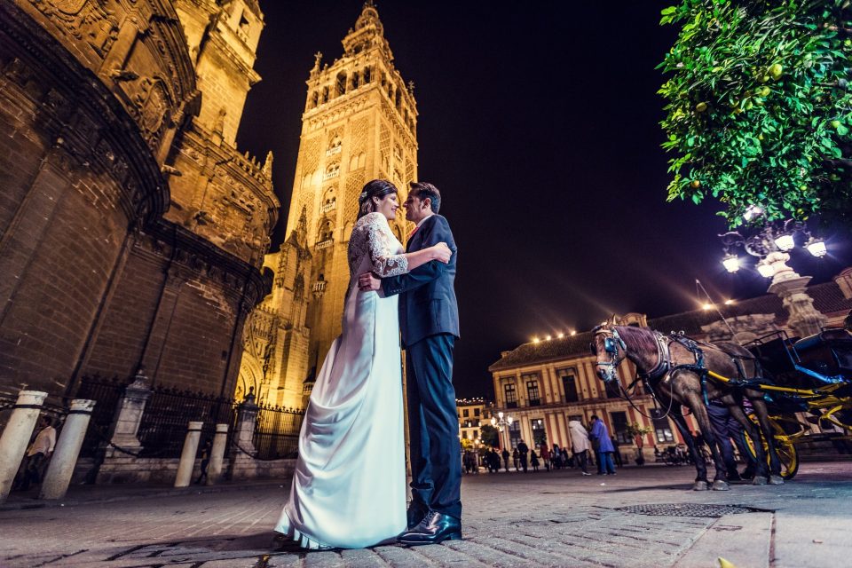 A couple in wedding attire embraces in front of an illuminated cathedral at night.