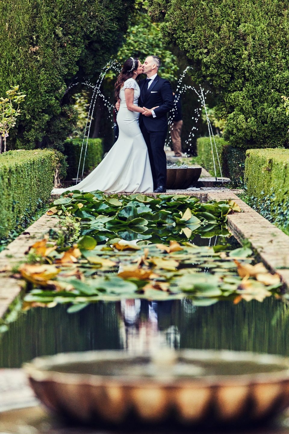 Una pareja vestida de boda se besa junto a una fuente en un jardín.