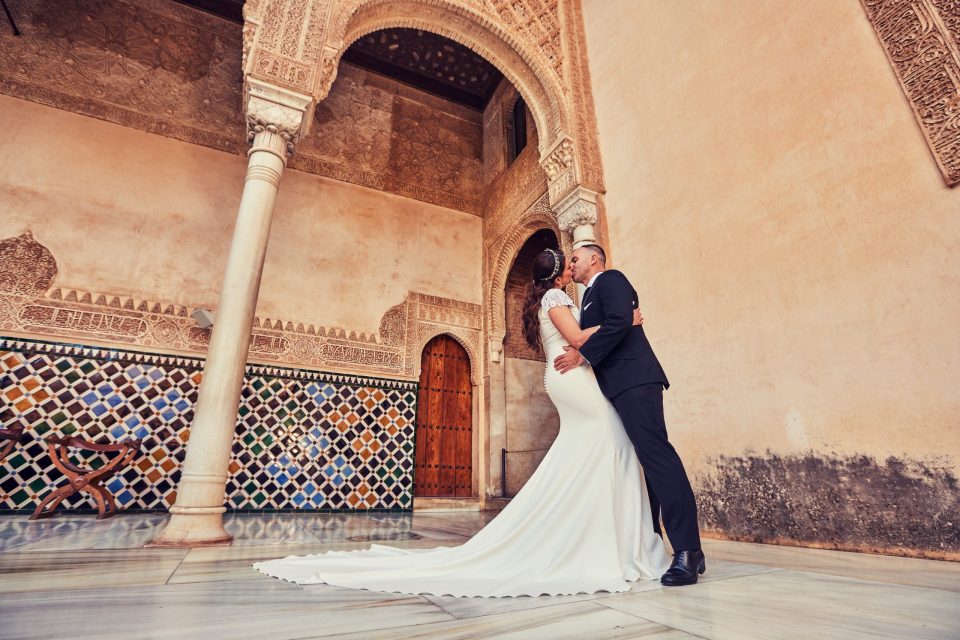 Una pareja vestida de boda se besa y abraza en un patio ornamentado.