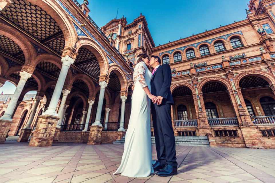 A couple in wedding attire kiss and hold hands in front of a historic brick building with large arches.