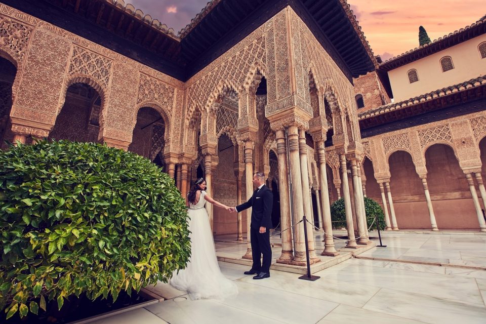 Una pareja vestida de boda se toma de la mano en un patio de arquitectura ornamentada, rodeado de arcos.