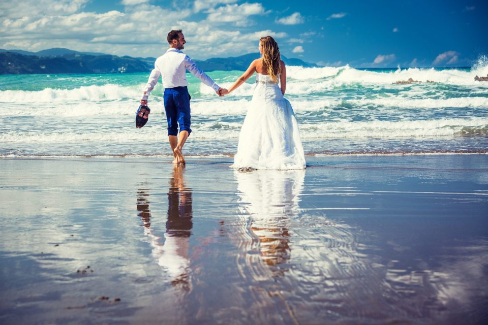 Una pareja vestida de novios camina de la mano por la orilla de una playa.
