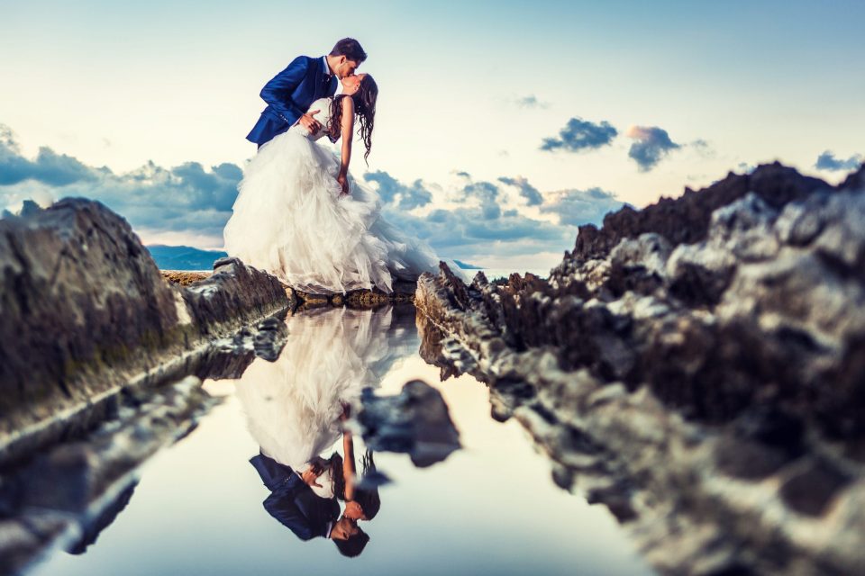 Una pareja vestida de novios se besa sobre unas rocas junto al mar.