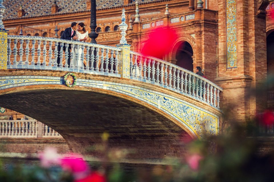 An elegantly dressed couple embrace and chat on an ornate bridge decorated with tiles and a balustrade.