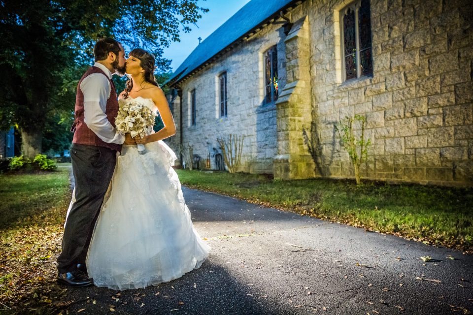 A couple dressed for their wedding kiss in front of a stone building with ornate windows.